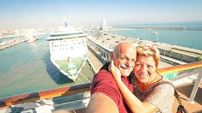 Elderly couple taking a selfie on a cruise ship deck with another large ship docked at a sunny harbor in the background.